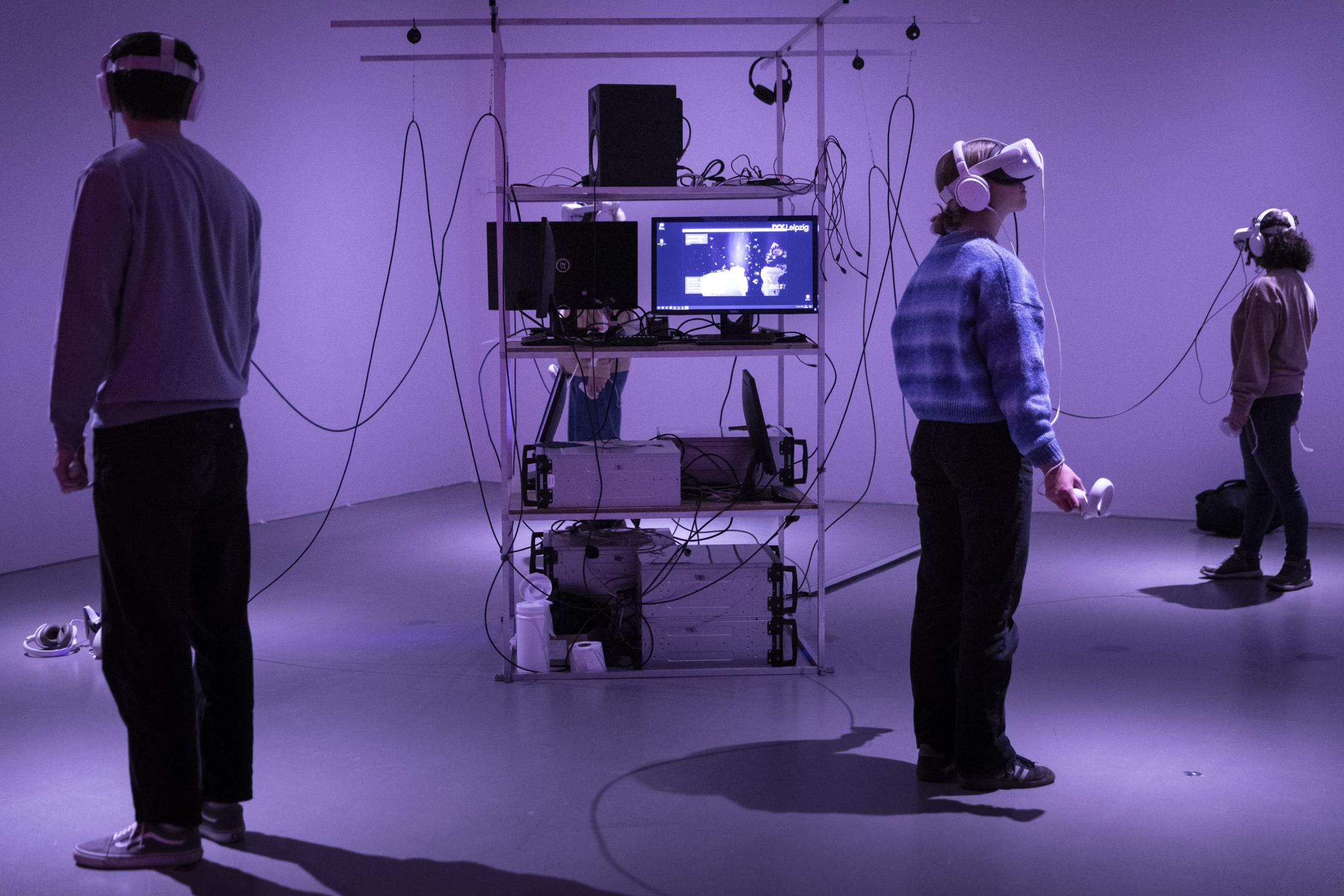 Drei Personen mit VR-Brillen stehen im Kreis um einen Turm aus digitalen Geräten. Sie blicken alle zur Seite.